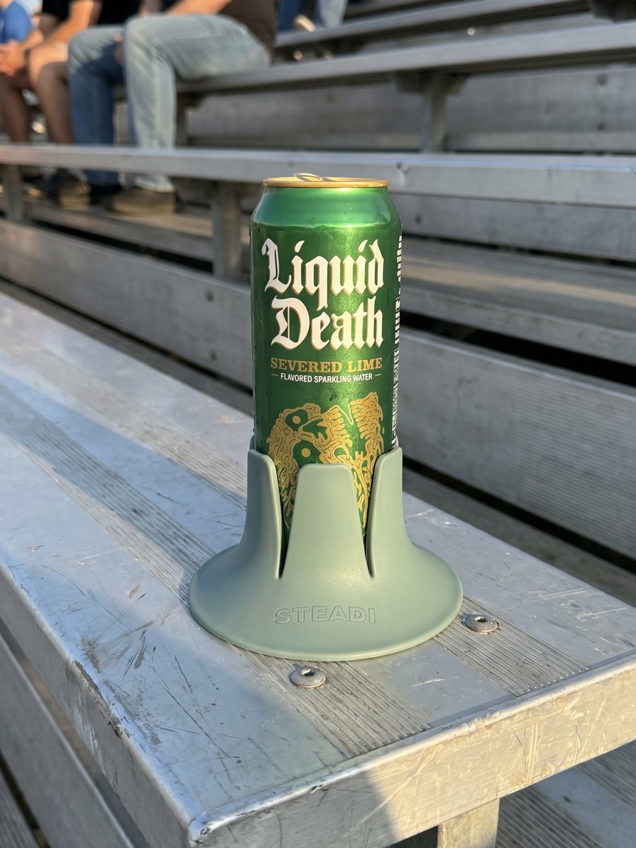 A sage Steadi holding a tall can on a scuffed aluminum bleacher plank, with empty rows and other spectators behind it.
