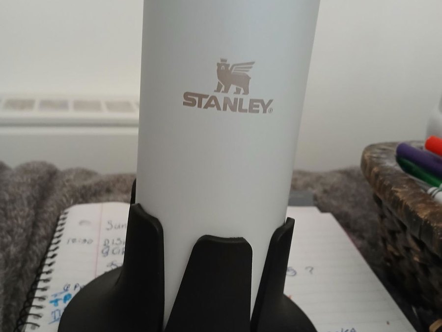 A white Stanley insulated tumbler seated in a black drink base standing on an open spiral planner covered in handwritten to-do notes, with a basket of pens alongside